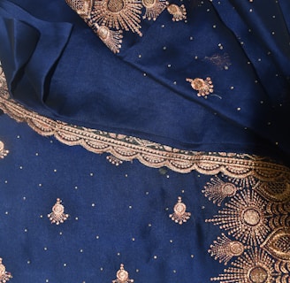 A rich blue fabric adorned with intricate gold embroidery, featuring floral and paisley patterns. The ornate designs are meticulously stitched with glittering gold thread, highlighting an elaborate and traditional textile craftsmanship.